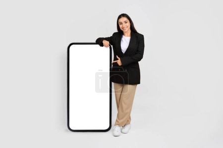 Foto de Friendly businesswoman in suit pointing at giant blank smartphone screen, perfect for advertising content, against a plain grey backdrop, mockup - Imagen libre de derechos