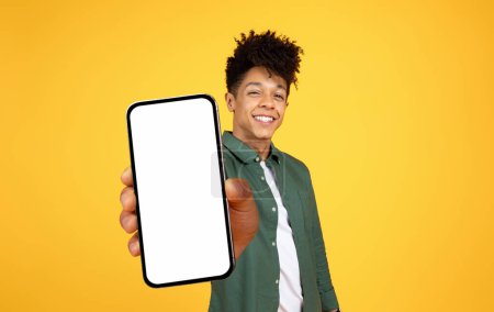 Téléchargez les photos : Cool cheerful handsome young black guy with stylish hairstyle showing big phone with white blank screen mockup copy space, isolated on yellow studio background. Mobile app - en image libre de droit