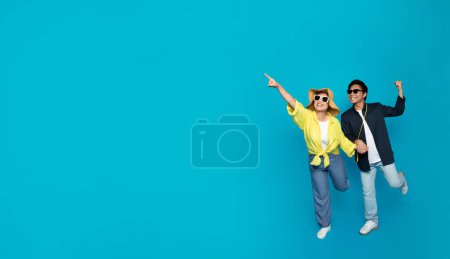 Téléchargez les photos : Ecstatic millennial Asian couple in summer attire are dancing and pointing joyfully, exuding happiness and carefree spirit on a bright turquoise background with ample copy space - en image libre de droit