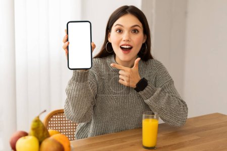 Foto de Amazed beautiful young woman showing nice mobile app for weight loss, sitting at table with fresh organic fruits and orange juice, holding smartphone with white blank screen, mockup, copy space - Imagen libre de derechos