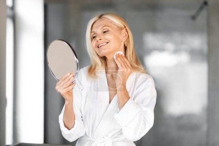 Foto de Smiling Senior Woman Cleansing Neck Skin With Cotton Pad And Looking In Mirror In Bathroom, Beautiful Elderly Female Wearing White Bathrobe Making Daily Beauty Routine At Home, Copy Space - Imagen libre de derechos