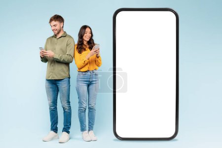 Téléchargez les photos : Man and woman using smartphones beside blank mockup screen, perfect for presenting apps or advertisements, with friendly, tech-savvy vibe against blue background - en image libre de droit