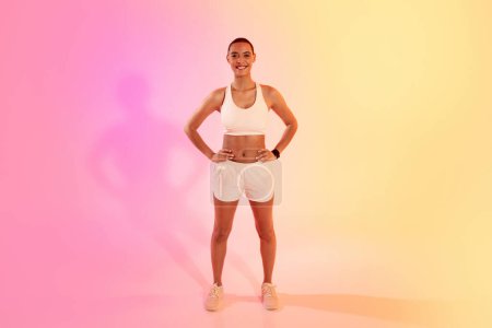 Téléchargez les photos : A confident female athlete stands with hands on hips in white sportswear, her shadow cast on a vibrant pink to yellow gradient background, exuding health and fitness. Sport, fitness and weight loss - en image libre de droit