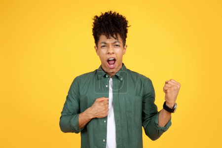 Foto de An exuberant emotional young african american man with curly hair punches the air with one fist, shouting in triumph, set against a stark yellow background - Imagen libre de derechos
