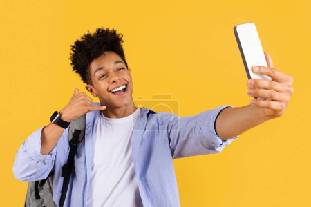 Photo for Exuberant male student making call me gesture while taking selfie with his smartphone, radiating happiness on vibrant yellow background - Royalty Free Image