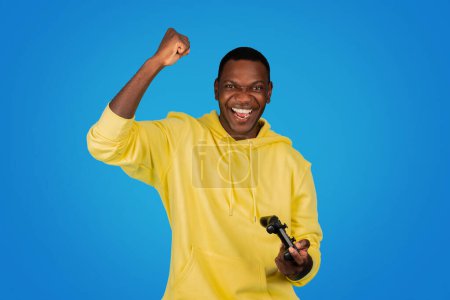 Foto de A victorious Black man in a yellow hoodie exudes triumph as he raises his fist in success while holding a game controller, set against a captivating blue studio backdrop - Imagen libre de derechos