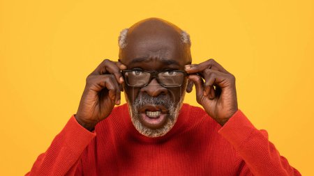 Téléchargez les photos : Surprised senior Black man adjusting his glasses with a puzzled expression, wearing a red sweater against a bright yellow background, signifying confusion or disbelief. - en image libre de droit