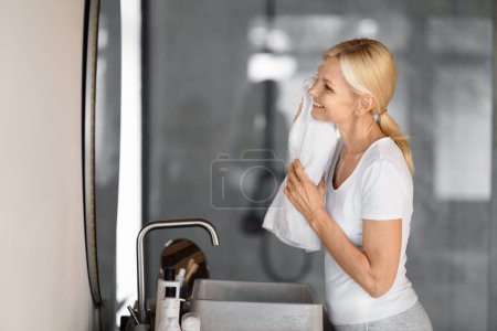Téléchargez les photos : Happy mature woman in casual clothes gently drying her face with soft white towel, smiling elderly female enjoying her skincare routine, standing in a modern, well-lit bathroom, side view - en image libre de droit