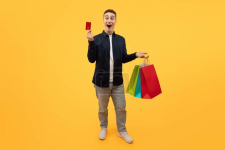 Photo for Emotional stylish guy shopaholic enjoying sale season black friday deal, showing his red bank credit card, carrying colorful paper bags purchases, yellow background. Shopping, consumerism - Royalty Free Image