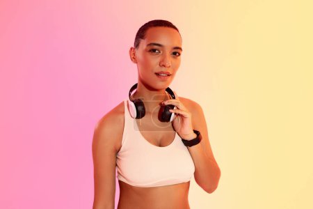 Téléchargez les photos : A serious woman with a shaved head wearing a white sports bra and a fitness tracker smiles while adjusting her headphones against a soft pink and yellow gradient background - en image libre de droit