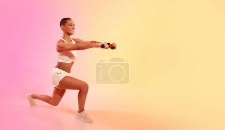 Téléchargez les photos : Exuberant young woman doing a lunge and holding weights, smiling during a workout session, dressed in a white sports top and shorts, with a soft pink and yellow gradient background - en image libre de droit