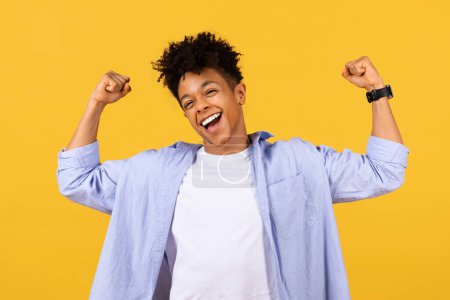 Photo for Joyful black male student with raised fists in victory pose smiling, exuding triumph and happiness, set against an uplifting yellow background - Royalty Free Image