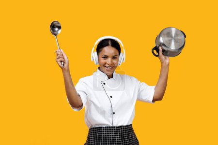 Téléchargez les photos : Happy black female chef enjoying music in headphones while holding ladle and pot, cheerful young african american cook woman having fun against bright yellow background, merging cooking with joy - en image libre de droit
