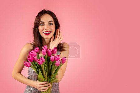 Photo for Vibrant young woman in a sparkling dress, cheerfully holding a bouquet of pink tulips and touching her face in surprise, against a pink background. Spring holiday celebration event - Royalty Free Image