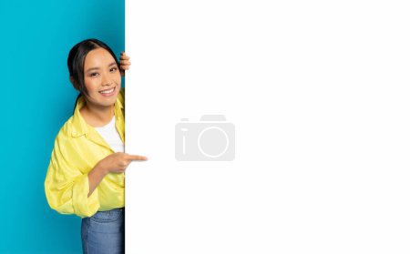 Photo for An Asian woman with a bright smile is peeking from behind a blank white vertical banner, playfully pointing towards it, ideal for customizable content on a turquoise background - Royalty Free Image