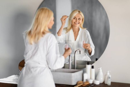 Téléchargez les photos : Elegant senior woman in white bathrobe applying facial serum, looking at her reflection in bathroom mirror, happy smiling elderly female enjoying her beauty routine at home, copy space - en image libre de droit