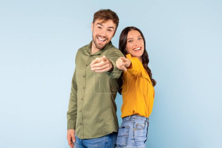 Photo for Smiling young spouses playfully pointing at the camera, their faces expressing warm invitation and friendliness, dressed in casual chic against light blue background - Royalty Free Image