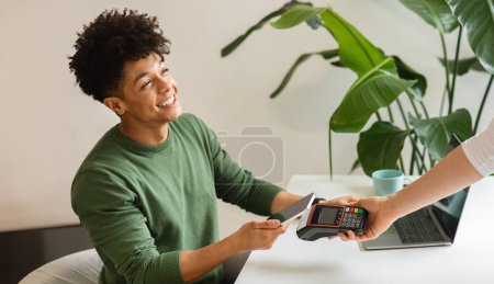 Foto de Positive stylish african american young man using smartphone while making contactless payment in a cafe. Cheerful black guy freelancer working at coffeeshop, paying for service with banking mobile app - Imagen libre de derechos
