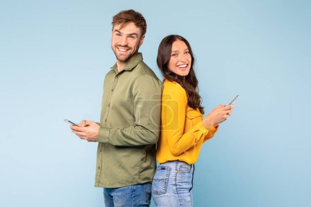 Téléchargez les photos : Happy young couple standing back-to-back, each using their own smartphone with joyful expressions, symbolizing individual digital engagement in social setting, against blue background - en image libre de droit