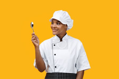 Téléchargez les photos : Smiling black female chef in uniform looking at spoon in her hand, african american cook woman suggesting hint of culinary mischief, standing against bright yellow background, copy space - en image libre de droit