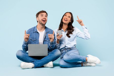 Téléchargez les photos : Enthusiastic young couple seated with laptop and smartphone, looking upwards with bright ideas, symbolizing moment of inspiration and creativity, against blue backdrop - en image libre de droit