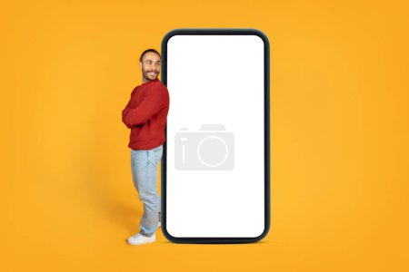 Téléchargez les photos : Curious cheerful stylish millennial man with arm crossed on chest looking at big phone with white blank screen and smiling, checking nice online offer, deal or new mobile app, orange background - en image libre de droit