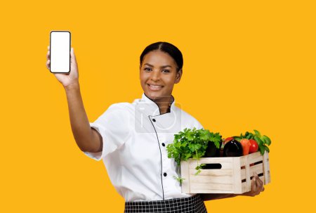 Foto de Grocery Market App. Cheerful Black Chef Woman Showing Smartphone With Empty Screen And Holding Crate With Fresh Organic Vegetables, Standing Over Yellow Studio Background. Mockup, Collage - Imagen libre de derechos