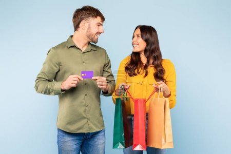 Foto de Happy man and woman enjoying shopping spree, man holding credit card and woman with multiple colorful shopping bags, sharing joyful moment on blue background - Imagen libre de derechos