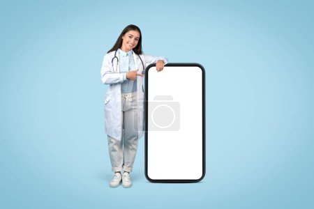 Photo for Confident young female doctor pointing to blank screen of smartphone, representing mobile health applications, on soothing blue background, mockup - Royalty Free Image