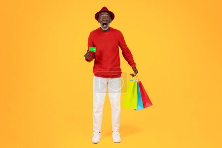 Foto de Glad surprised senior african american man shopaholic in hat with open mouth and many bags with purchases, show credit card, isolated on orange studio background. Shopping, sale and money - Imagen libre de derechos