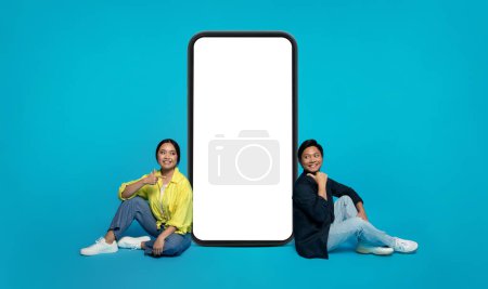 Téléchargez les photos : Smiling Asian millennial couple sitting on the floor with a giant smartphone between them, giving thumbs up, implying a positive review or endorsement on a turquoise background - en image libre de droit
