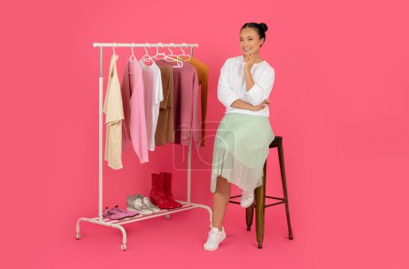 Photo for Beautiful Young Asian Fashion Stylist Woman Sitting On Chair Near Clothing Rack, Happy Korean Female Thinking About Trends And Shopping Sales, Posing On Pink Studio Background, Copy Space - Royalty Free Image