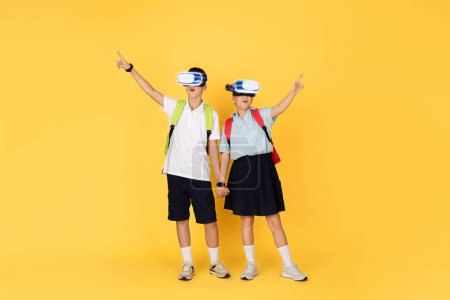 Photo for Two enthusiastic school students with backpacks hold hands while experiencing virtual reality through headsets, gesturing in amazement against a vivid yellow background - Royalty Free Image