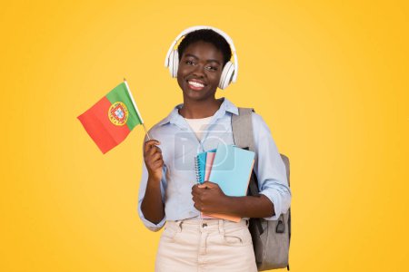 Téléchargez les photos : Happy teen african american lady student in wireless headphones, with books, flag of Portugal, listen lesson, isolated on yellow studio background. Study and education exchange, learn Portuguese - en image libre de droit