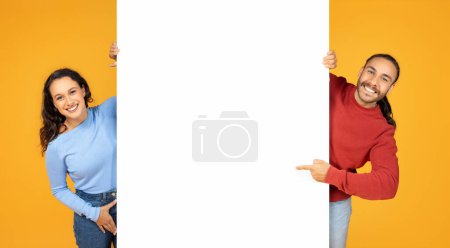 Téléchargez les photos : Happy millennial couple cheerful european man and woman showing white blank advertising board between them, pointing at placard with mockup copy space, orange background - en image libre de droit