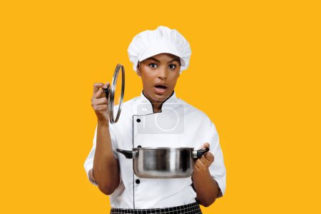 Photo for Surprised black female chef lifting the lid off pot and looking at camera, african american cook woman capturing her unexpected reaction to the dish, standing against bright yellow background - Royalty Free Image