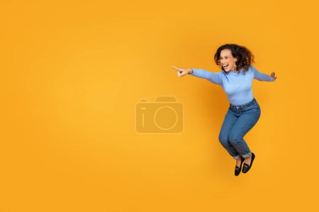 Photo for Great offer. Cheerful excited young woman wearing comfy casual clothing moving, jumping in the air, have fun alone on yellow orange wall background, feeling happy, pointing at copy space, full length - Royalty Free Image