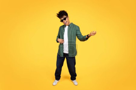 Téléchargez les photos : Stylish young black guy wearing sunglasses and casual clothing playing imaginary guitar isolated on yellow studio background. Cool african american millennial man rockstar, copy space, full length - en image libre de droit