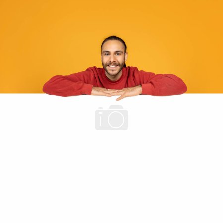 Téléchargez les photos : Positive handsome young guy hipster leaning on blank white board for text or advertisement, copy space, mockup, isolated on orange studio background. Ad, offer, deal - en image libre de droit