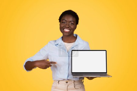 Photo for Glad young african american woman student in casual point finger at computer laptop with empty screen, isolated on yellow studio background. Study and education app, attention website for lesson - Royalty Free Image