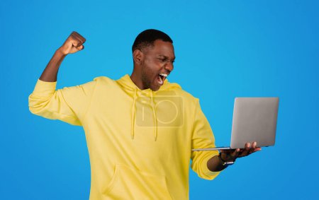 Foto de An exuberant Black man in a yellow hoodie raises his fist in victory as he looks at a laptop, his face expressing triumphant joy against a bright blue studio backdrop - Imagen libre de derechos