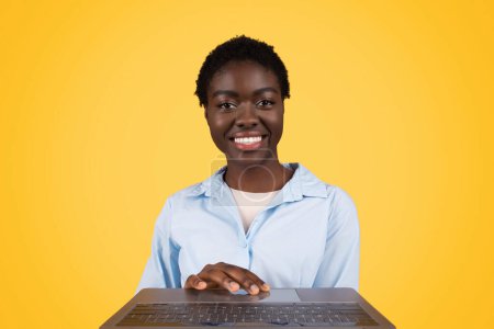 Téléchargez les photos : Happy young african american lady student in casual typing on computer keyboard, enjoy online lesson, chat, isolated on yellow studio background. Study and education, app, social networks - en image libre de droit