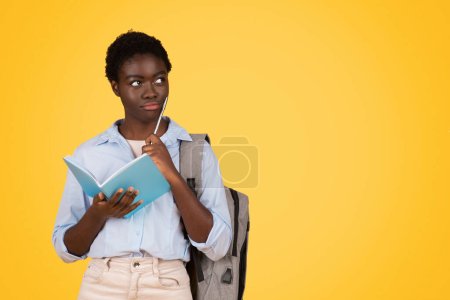 Téléchargez les photos : Pensive african american teen lady student, think, dream, make writes in diary, isolated on yellow background, studio. Education planning, study, homework and lesson project - en image libre de droit
