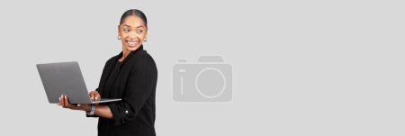 Foto de Una alegre mujer de negocios afroamericana sostiene una computadora portátil con una mano mientras sonríe y mira hacia un lado, retratando una sensación de facilidad y experiencia, aislada en un fondo gris de estudio - Imagen libre de derechos