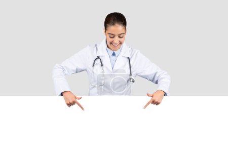 Téléchargez les photos : Une dame médecin européenne souriante en manteau blanc avec un stéthoscope pointe vers le bas vers une bannière vide, parfaite pour la publicité des services de santé ou d'information, de vente, de conseils - en image libre de droit