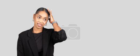 Téléchargez les photos : Une jeune femme afro-américaine professionnelle réfléchie dans un blazer noir a l'air surprise, main sur la tête, avec un sourire subtil, isolée sur fond de studio gris, panorama - en image libre de droit