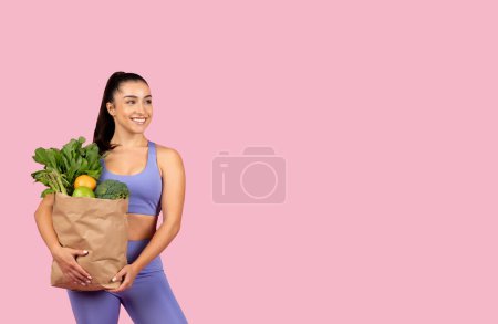 Foto de Mujer de corte europeo feliz en ropa deportiva con bolsa llena de frutas y verduras frescas mirando el espacio libre aislado en el fondo del estudio rosa. Pérdida de peso y cuidado corporal, dieta - Imagen libre de derechos