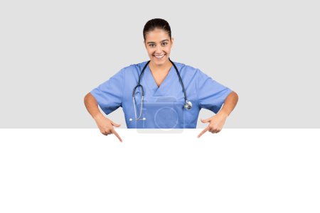 Téléchargez les photos : Une dame médecin européenne souriante en blouse bleue se penche sur une bannière vierge, pointant vers le bas où des informations sur les soins de santé ou du contenu promotionnel peuvent être ajoutés, isolée sur fond de studio gris - en image libre de droit