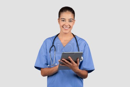 Téléchargez les photos : Jeune infirmière souriante et technicienne en blouse bleue, utilisant une tablette numérique, symbolisant la gestion moderne des soins de santé et de l'information, avec un stéthoscope autour du cou - en image libre de droit