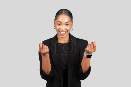 Téléchargez les photos : Radieuse jeune femme d'affaires noire en blazer fait un mini geste de cœur avec ses mains, souriant joyeusement, tenir de l'espace libre, isolé sur fond de studio gris. Travail, plaisir, entreprise - en image libre de droit